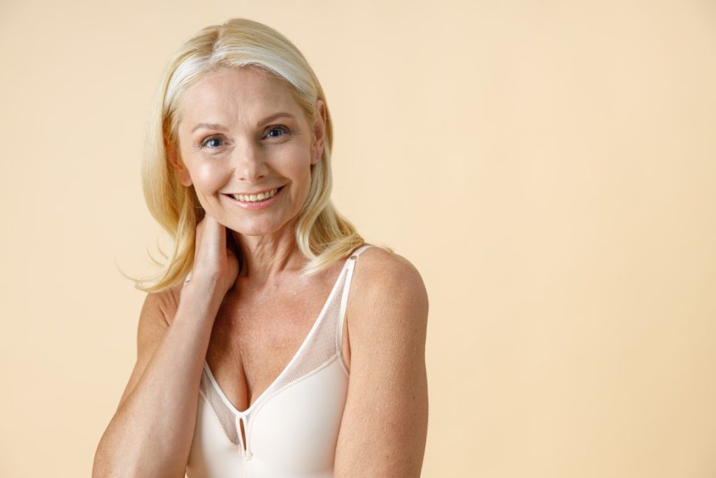 portrait of charming mature woman with blonde hair in white underwear smiling at camera, posing isolated over beige background